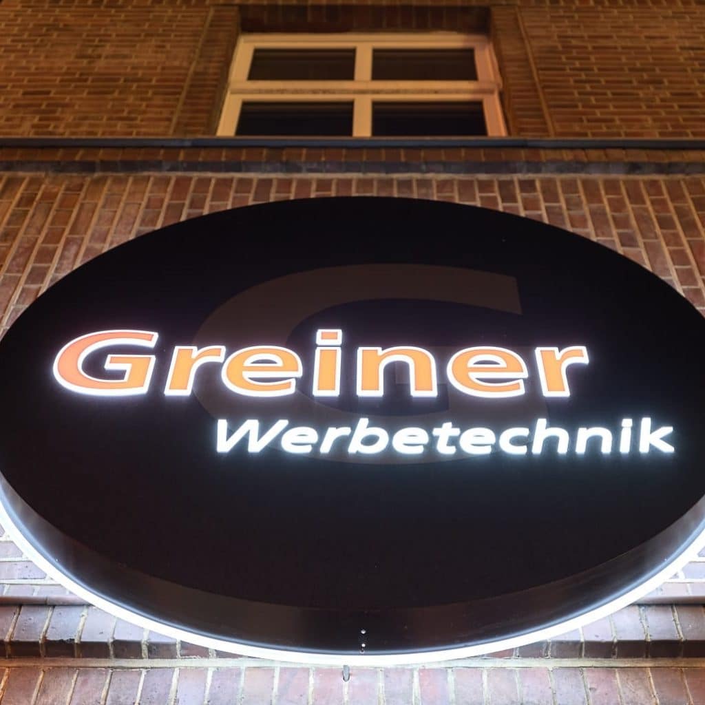 Leuchtendes rundes Firmenschild mit der Aufschrift "Greiner Werbetechnik" vor einer Backsteinwand und Fenstern im Hintergrund.
