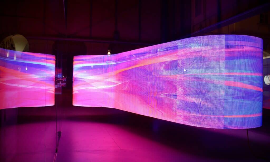 Gebogene, leuchtende LED-Anzeige mit abstrakten, farbenfrohen Lichtmustern in Pink, Lila und Blau, die sich in einer Glaswand spiegelt.