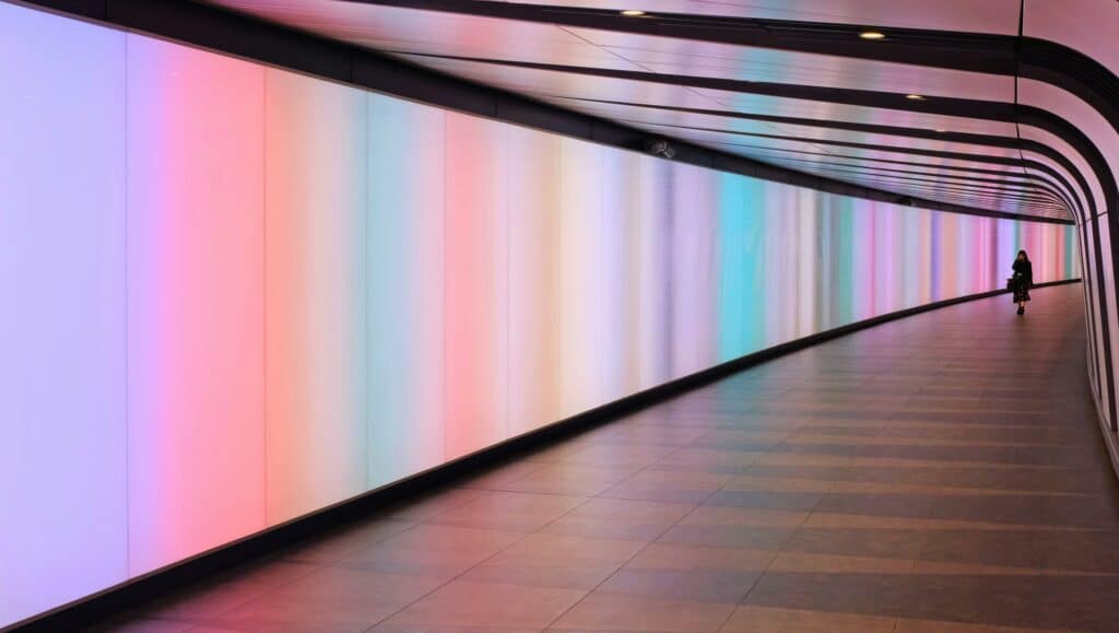 Geschwungener Tunnel mit bunten, leuchtenden Wandpanelen in Pastellfarben und einer einzelnen Person, die in der Ferne geht.