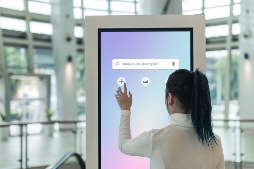 Eine Frau mit dunklem Pferdeschwanz bedient einen großen interaktiven Touchscreen mit einer Suchleiste und Symbolen in einer modernen, hellen Umgebung.