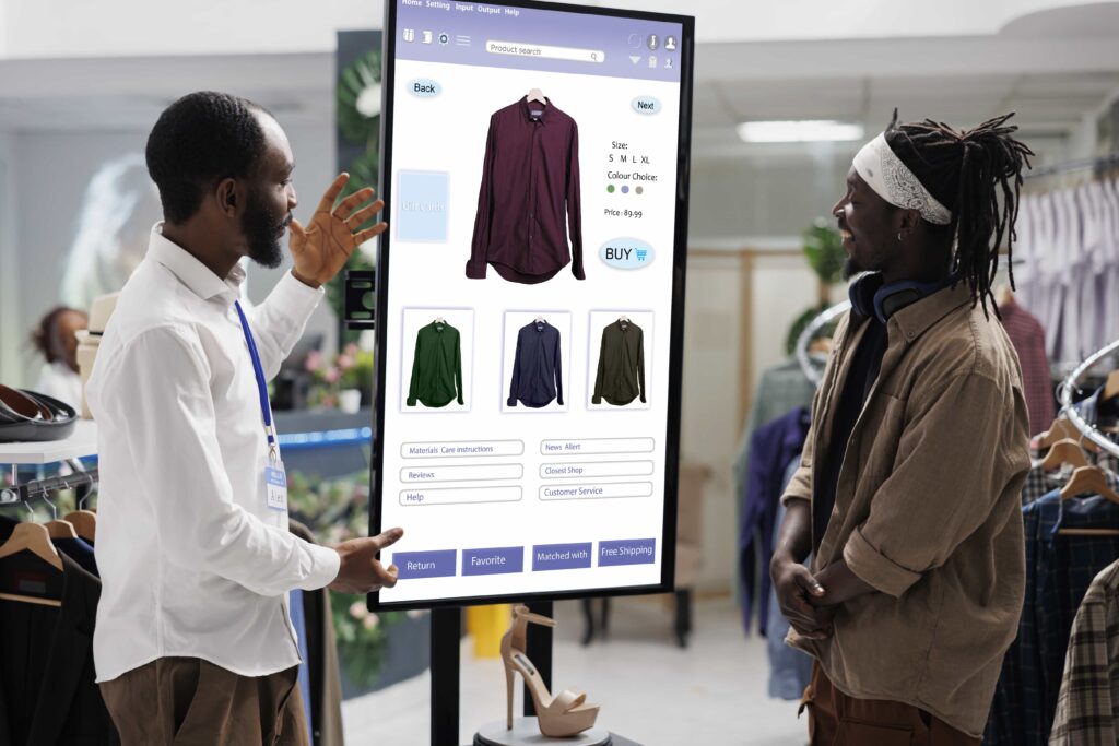 Zwei Männer stehen vor einem großen Touchscreen in einem Bekleidungsgeschäft, auf dem eine Online-Shop-Oberfläche mit Hemden und Bestelloptionen angezeigt wird.